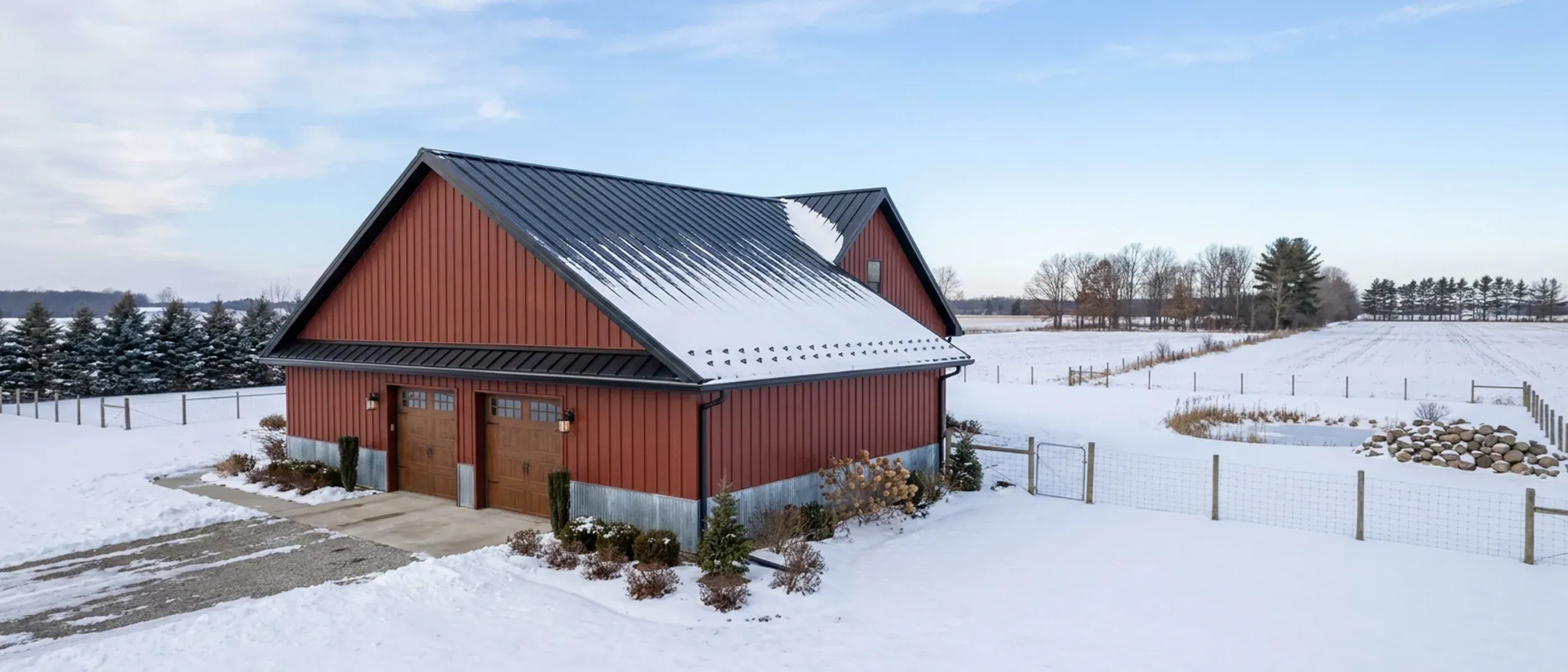 A completed barndominium in winter viewed from a low angle, showing its standing seam roof with snow guards, wall panels, gutters and wall lights. A completed barndominium in winter viewed from a low angle, showing its standing seam roof with snow guards, wall panels, gutters and wall lights.